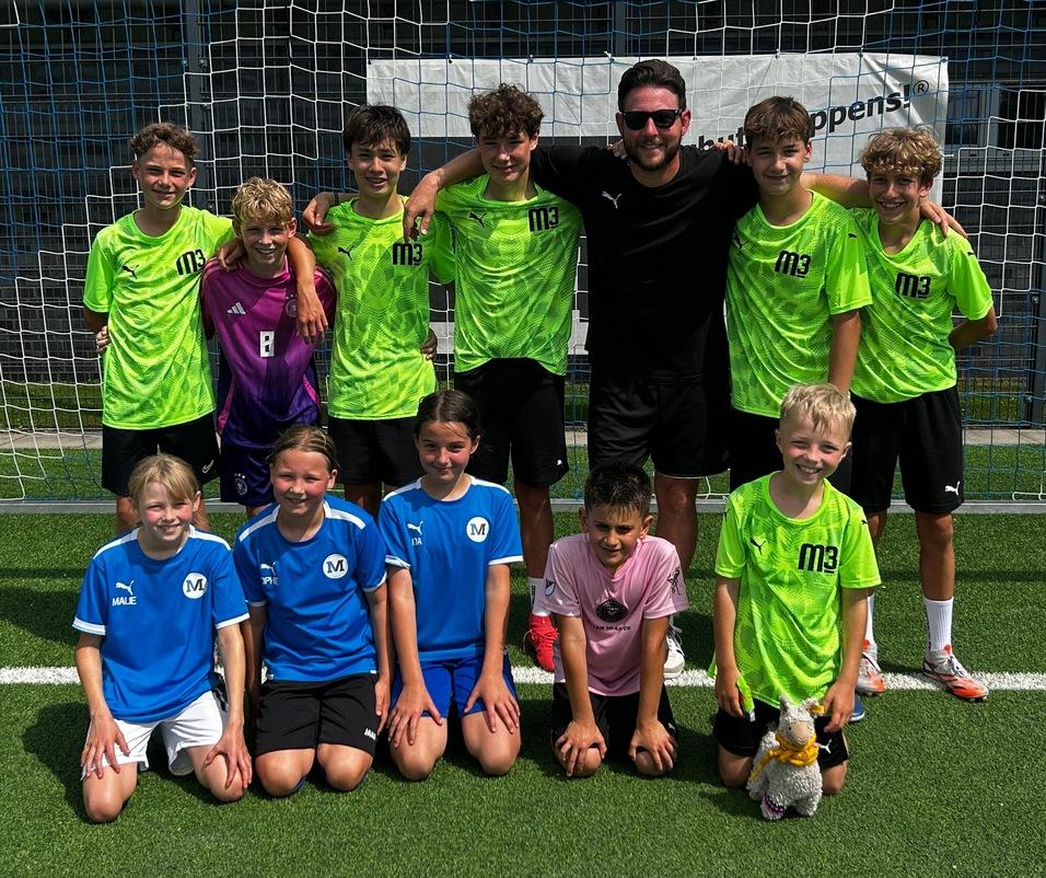 Gruppe von zehn Jugendlichen in Fußballtrikots, posierend vor einem Tor auf einem Rasenplatz.